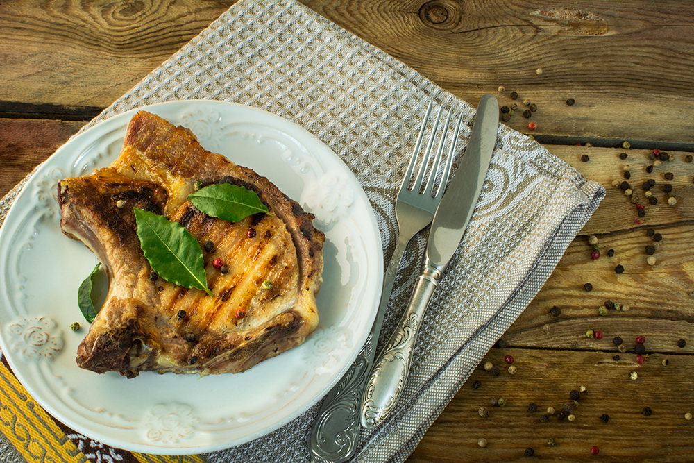 fried steak with green laurel leaves on white plate, knife, fork, linen napkin and grains of pepper wooden background